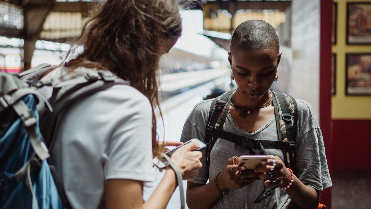 Dos viajeras con mochila revisando sus teléfonos en una estación de tren