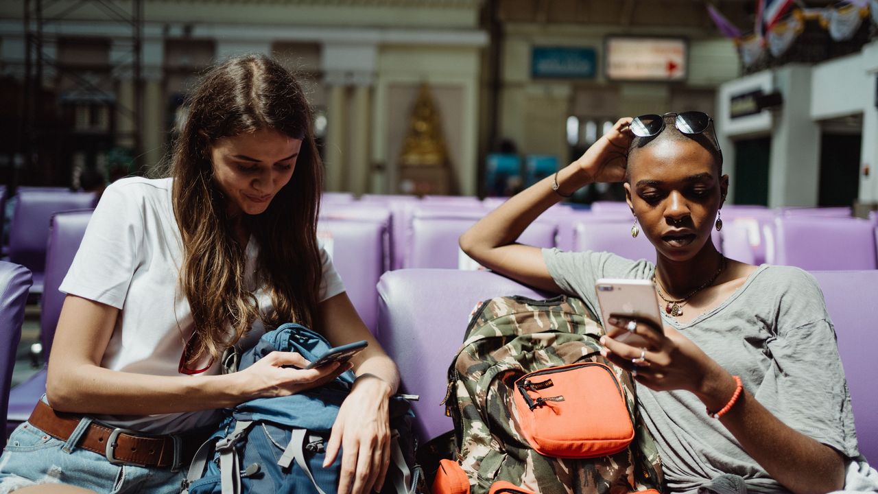 Dos viajeras con mochila miran sus teléfonos en una estación de tren