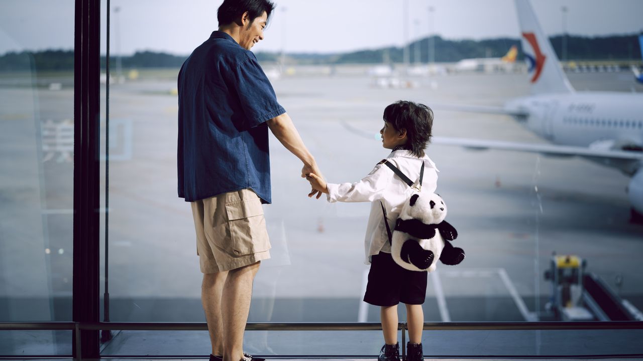 Padre e hijo tomados de la mano junto a la ventana de un aeropuerto mirando los aviones