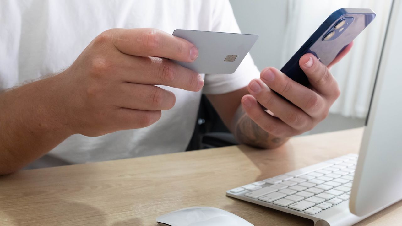 Manos que sostienen una tarjeta bancaria y un teléfono en un escritorio, a punto de pagar en línea