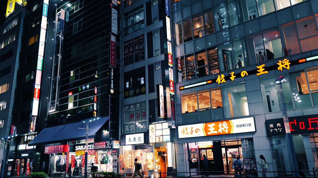 People walking along a Tokyo street at night under lit shop signs and tall office buildings