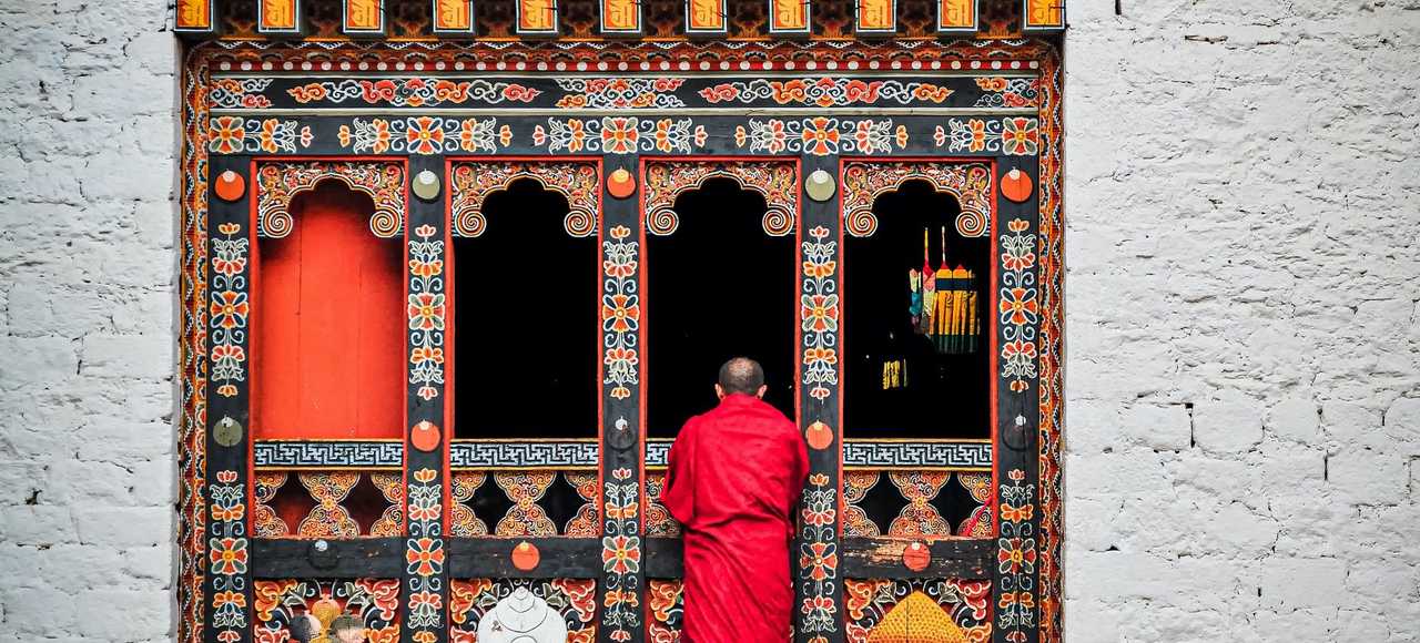 A Buddhist monk in red robes standing at an ornately decorated temple doorway in Bhutan