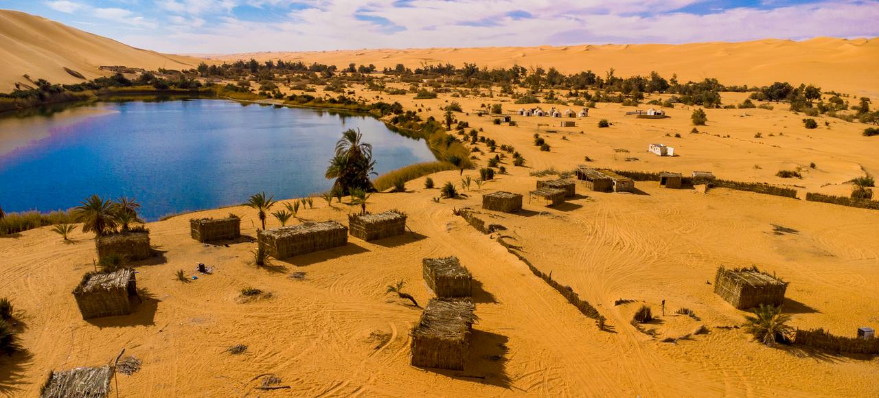 Chad: Aerial view of tranquil oasis surrounded by Sahara desert dunes and rustic huts