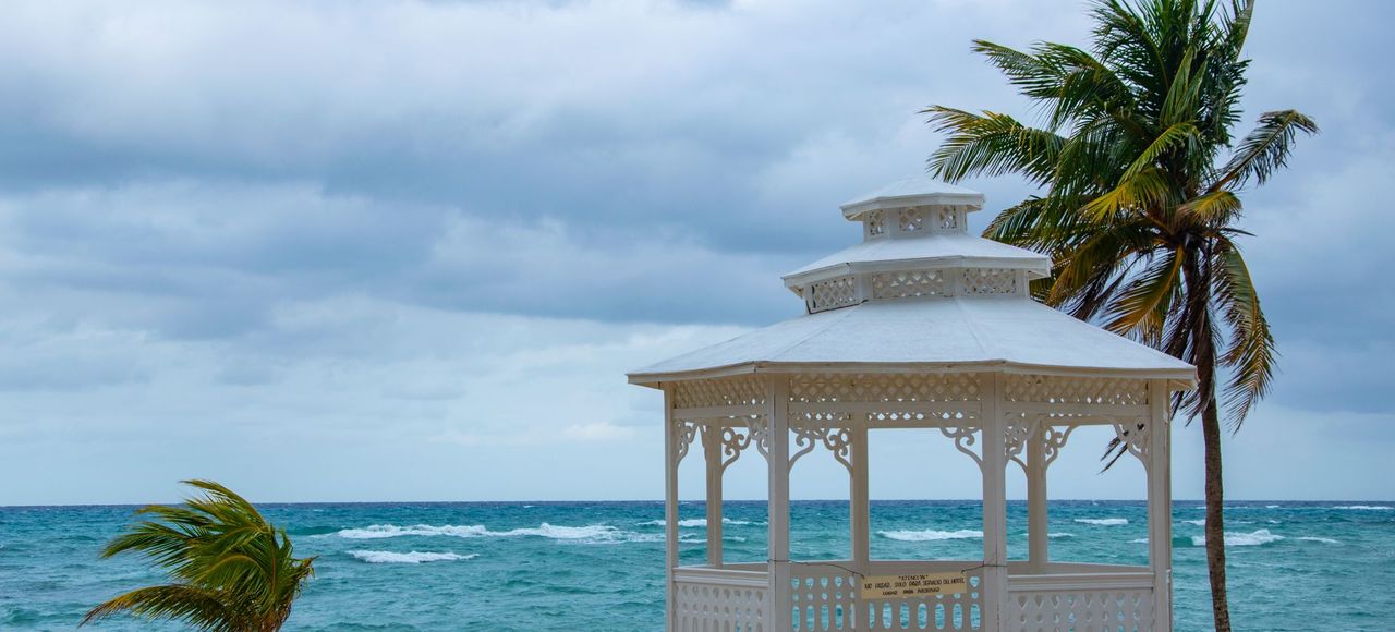 Jamaica: A serene tropical beach scene featuring a white gazebo, palm trees, and the ocean under a cloudy sky