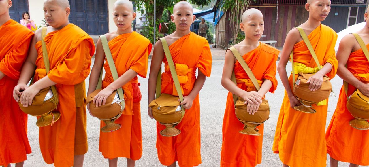 Laos: A group of young monks dressed in orange robes standing outdoors, holding alms bowls
