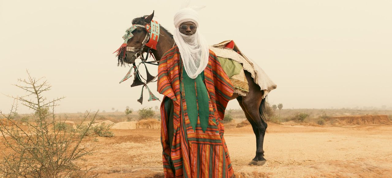 Niger: A Tuareg nomad in vibrant attire standing beside a camel in a vast desert landscape