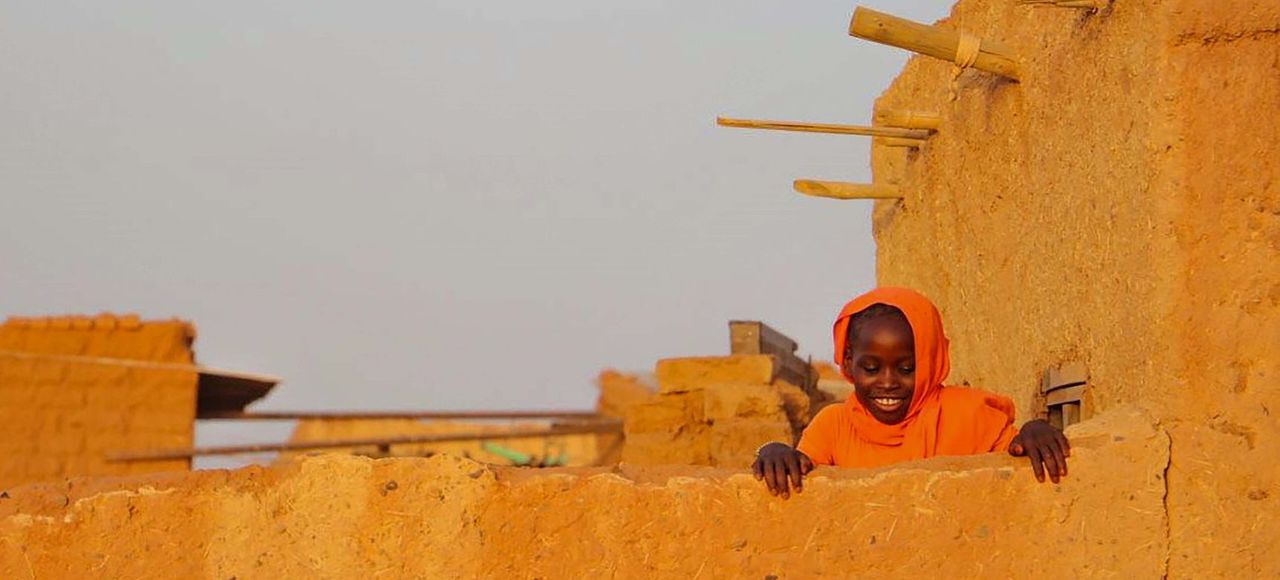 A joyful child in bright orange attire smiles over a rustic stone wall in Khartoum, Sudan