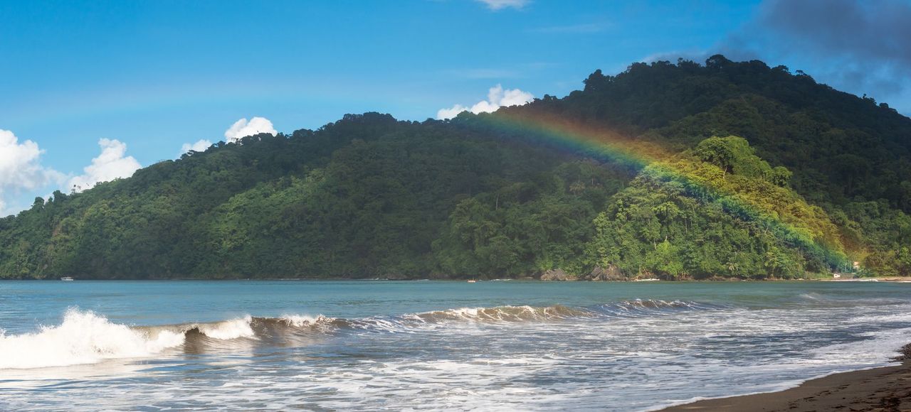 Vibrant rainbow stretches over a lush green hill by a sandy beach in Trinidad and Tobago
